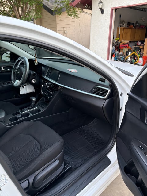 White car with open door showing black interior, steering wheel, and dashboard, parked in driveway with garage visible in background