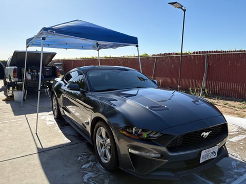 Black Ford Mustang parked under a blue canopy tent at a car wash facility with metal fence in background