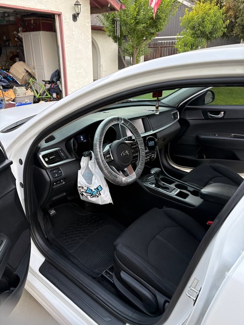 Interior of a white car with black seats, steering wheel, and dashboard visible with an open drivers side door in a residential driveway