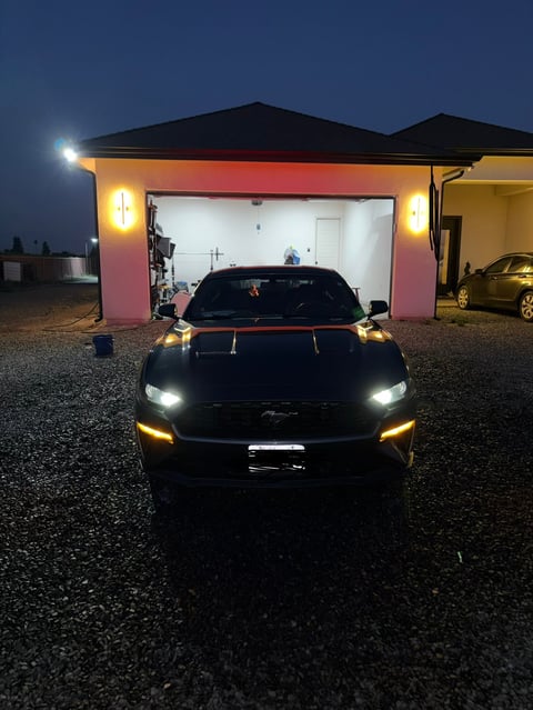 Black muscle car with glowing headlights and yellow accents parked in front of illuminated garage at dusk