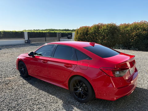 Red sedan parked on gravel with metal fence and trees in background
