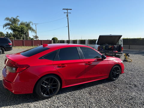 Bright red sedan parked on gravel lot with industrial buildings and utility poles in background