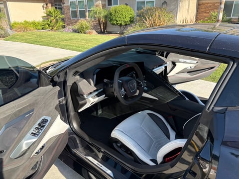 Black sports car with white and black interior, doors open, parked in driveway with houses visible in background