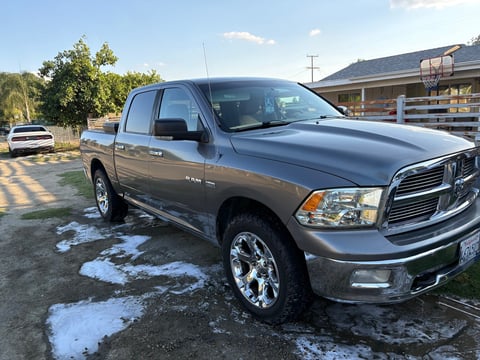 Silver Dodge Ram pickup truck parked in a driveway with snow patches, mountains and buildings visible in the background