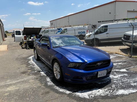 Blue Ford Mustang coupe parked in a lot with service vehicles and industrial building in background