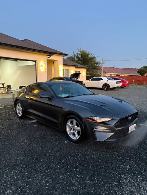 Black Ford Mustang parked in driveway in front of yellow house with other vehicles visible